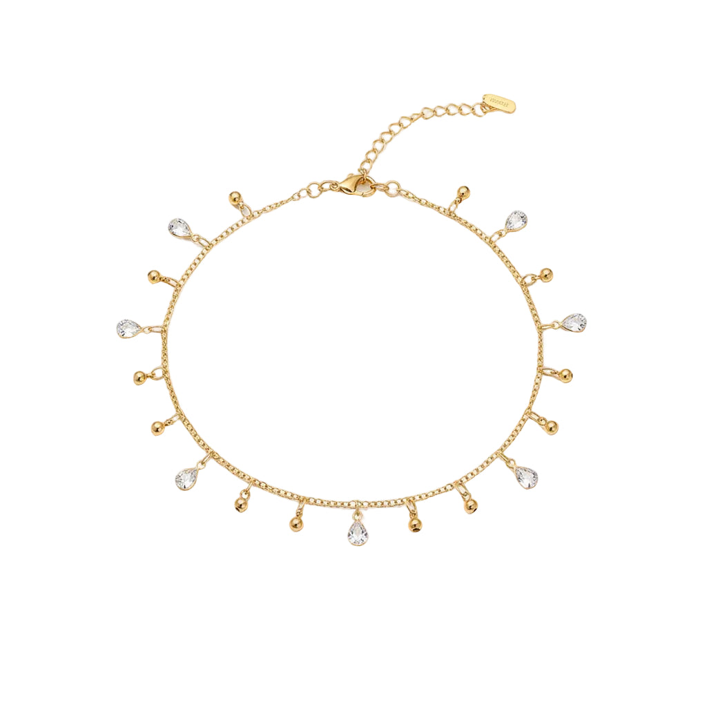 Gold anklet with small droplet charms on a foot against a sandy background